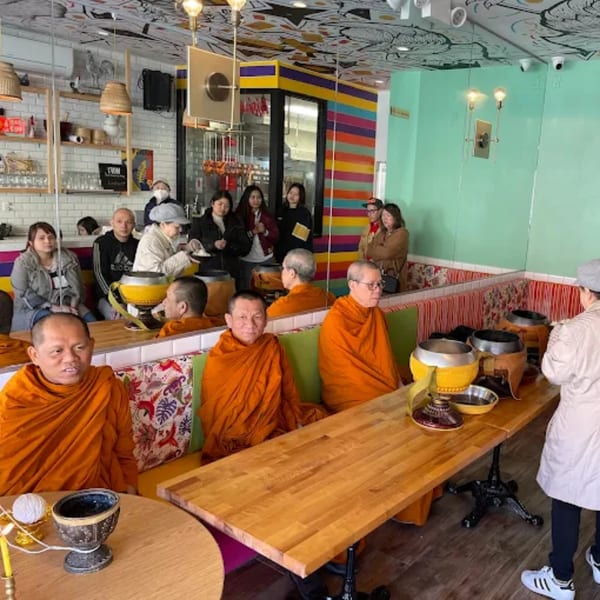 Monks in orange robes seated at tables with alms bowls, surrounded by people in a colorful Thai restaurant.
