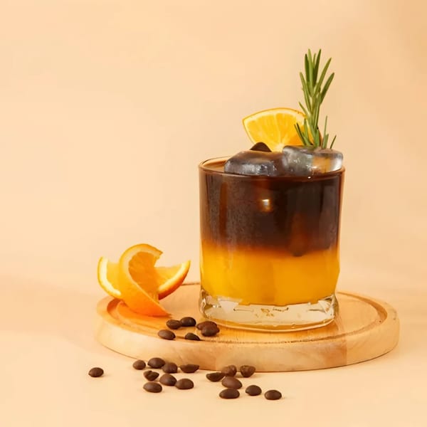 A glass of iced coffee with orange juice, garnished with an orange slice and rosemary.