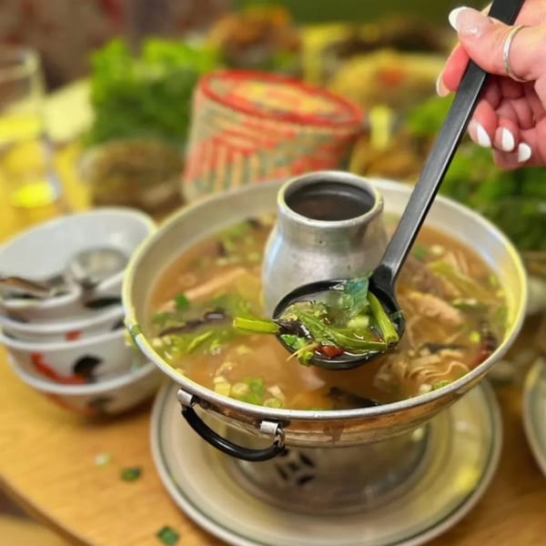 A hand spoons green vegetables from a large pot of Thai soup, with stacked bowls nearby.