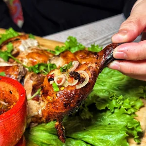 Hand holding a grilled chicken drumstick topped with onions and herbs, with lettuce and sauce.