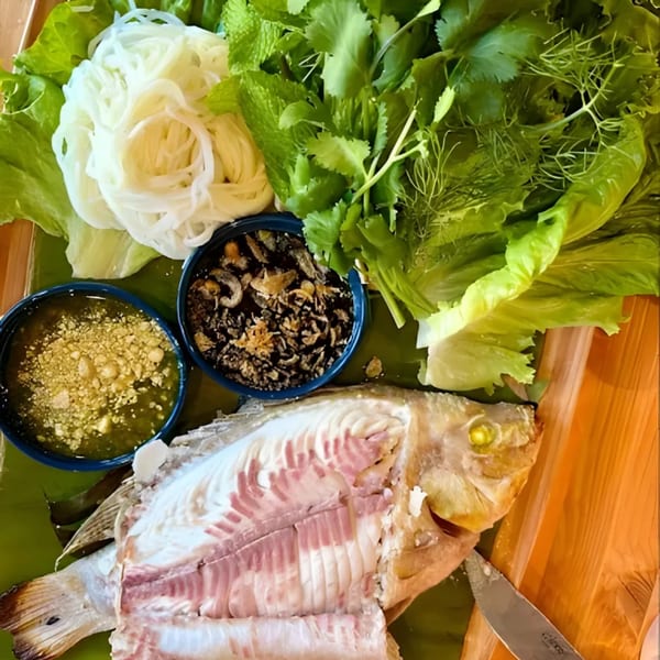 Whole grilled fish with herbs, noodles, and two dipping sauces on a wooden board.